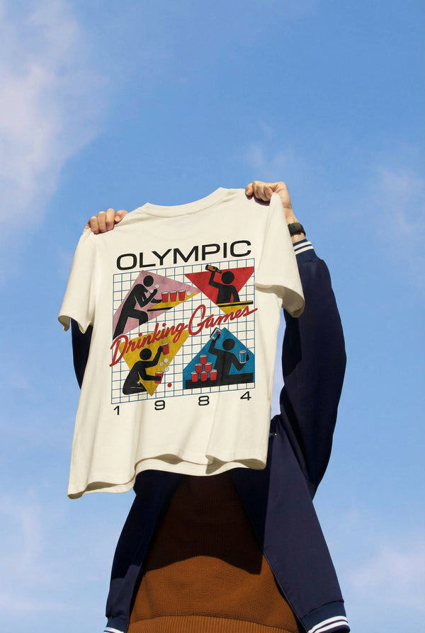 Person holding a t-shirt with 'Olympic Drinking Games 1984' graphic against a blue sky.
